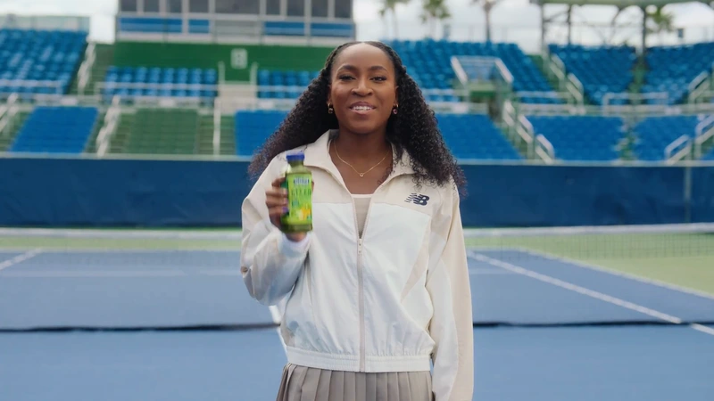 Coco Gauff holding a Naked Smoothie bottle while wearing a white zip-up jacket in a blue tennis stadium.