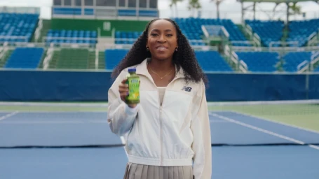 Coco Gauff holding a Naked Smoothie bottle while wearing a white zip-up jacket in a blue tennis stadium.