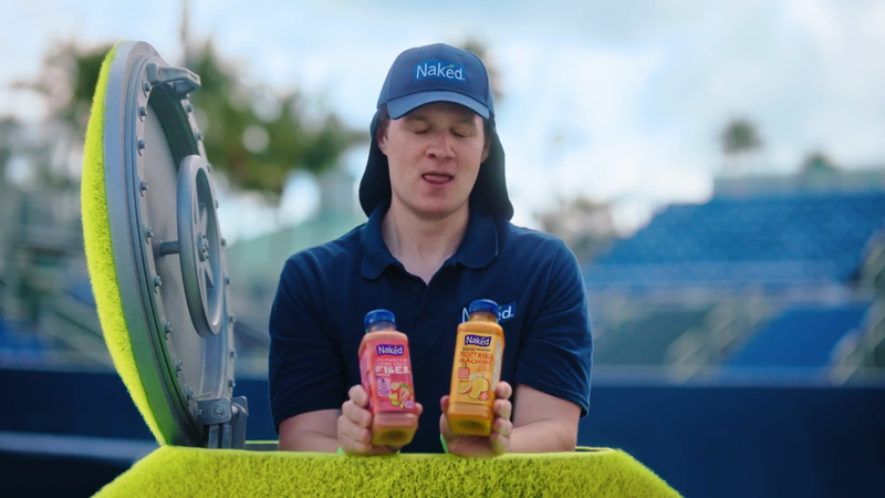 Man in a tennis umpire chair holds two Naked Juice bottles at an outdoor tennis court, promoting the brand's fruit smoothies.