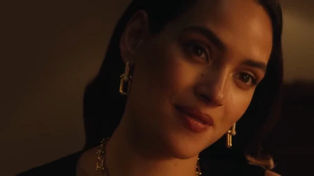 A woman wearing a gold chain necklace and earrings sits across from a man in a softly lit apartment setting for Tiffany and Co.