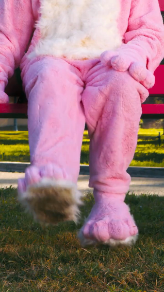 Person in a pink bunny costume sits on a park bench on grass, close-up of the legs and feet of the costume.