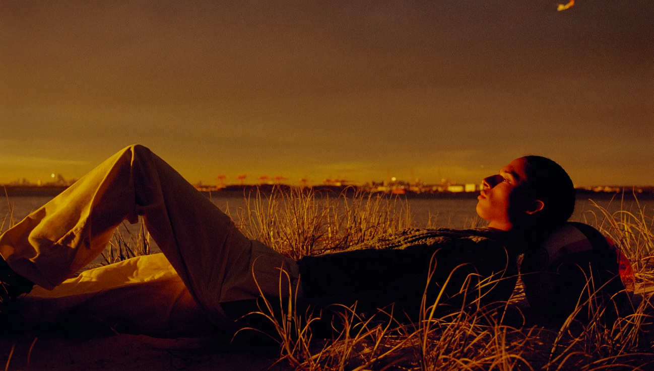 Person relaxes on grassy dunes at sunset, gazing at airplane in sky over water and distant urban skyline in golden hour lighting.