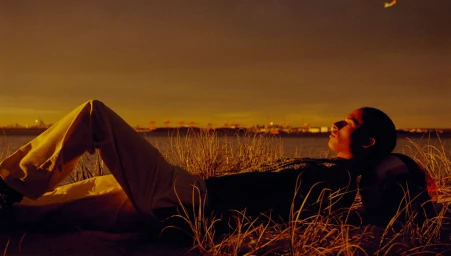 Man lying back in coastal grass at sunset, wearing light trousers and a dark sweater, with a distant body of water in the background.