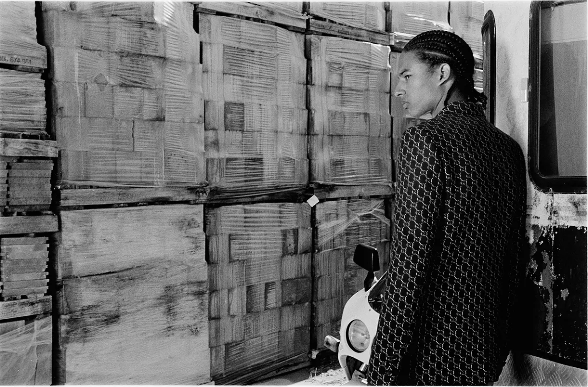 Adult male with cornrow braids poses next to a motorcycle and pallets wrapped in plastic in an indoor warehouse-like setting in monochrome.
