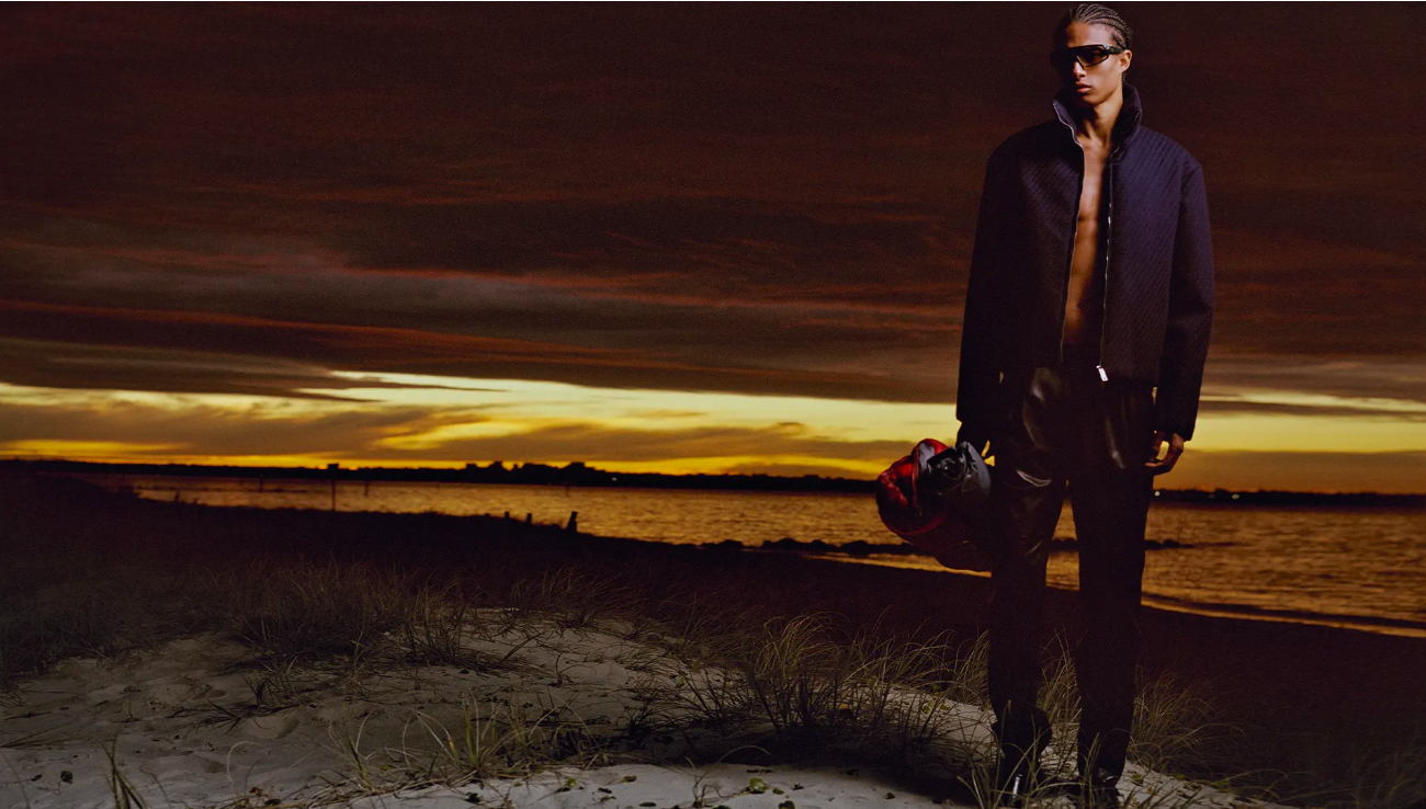 Man stands on a sandy beach at sunset, wearing a jacket and sunglasses, holding a helmet, with a dark cloudy sky and water background.