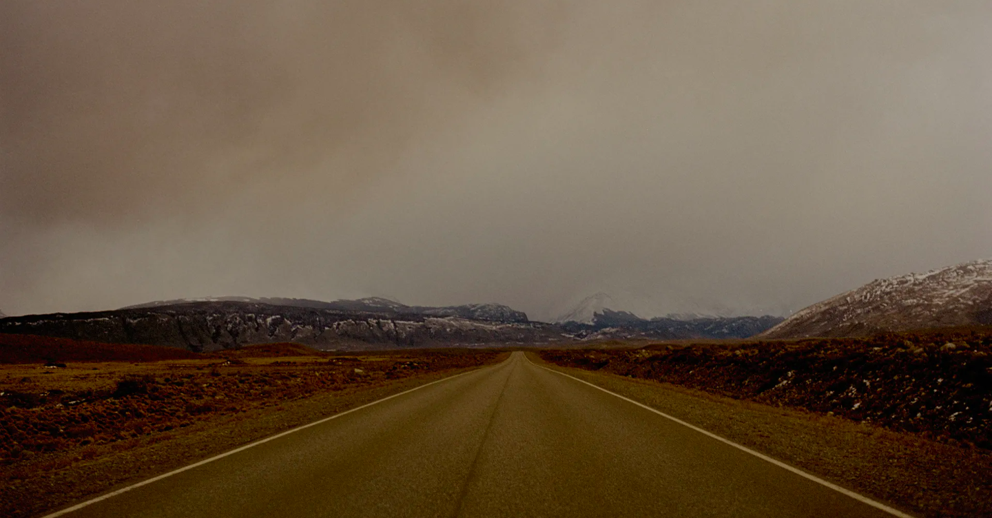 Asphalt road stretches to snow-dusted mountains under a cloudy sky in a rural landscape, creating a moody, desolate scene.