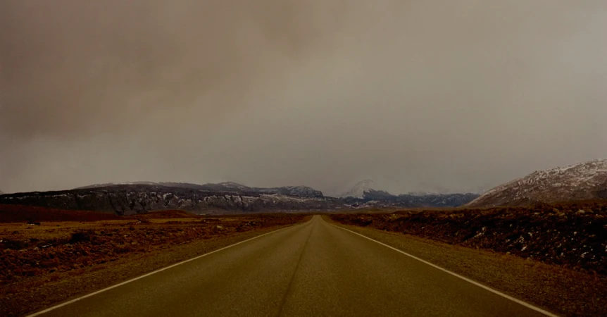Asphalt road stretches to snow-dusted mountains under a cloudy sky in a rural landscape, creating a moody, desolate scene.