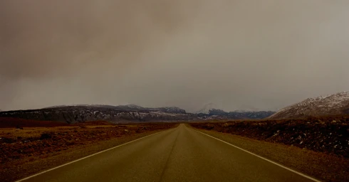 An empty road stretches towards a vast mountainous landscape under a heavy, overcast sky, photographed by Jake Terrey.
