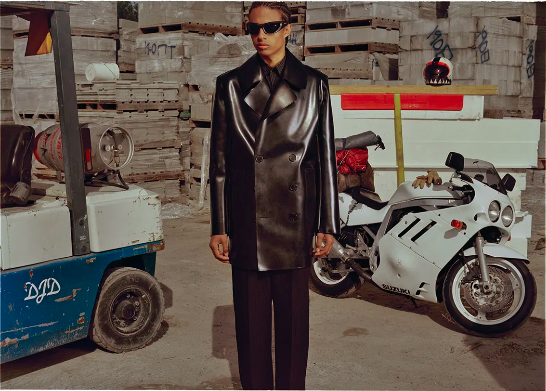 A stylish adult male poses in a black leather trench coat and sunglasses, with a motorcycle and industrial background.