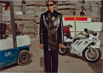 Man wearing a black leather coat and sunglasses standing outdoors near a white motorcycle and a blue forklift.