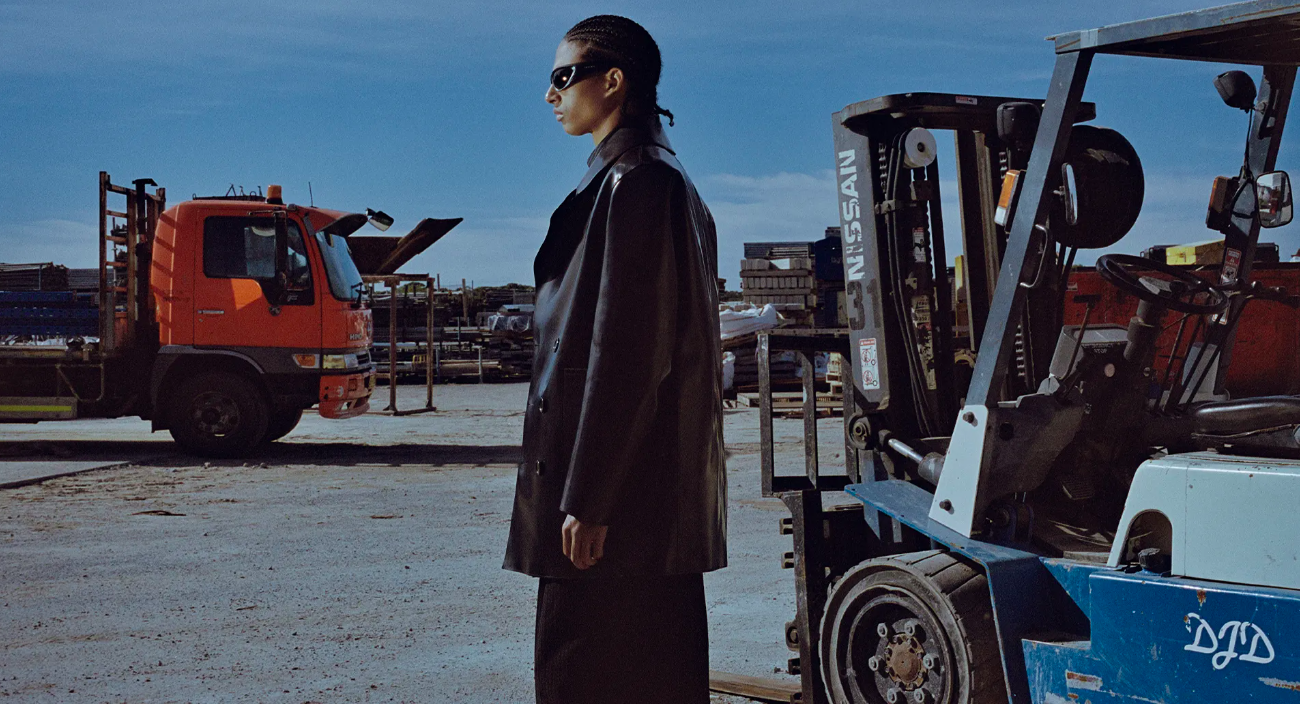 Side view of adult in a black leather jacket and sunglasses, standing in a lot with a forklift and an orange truck under blue skies.