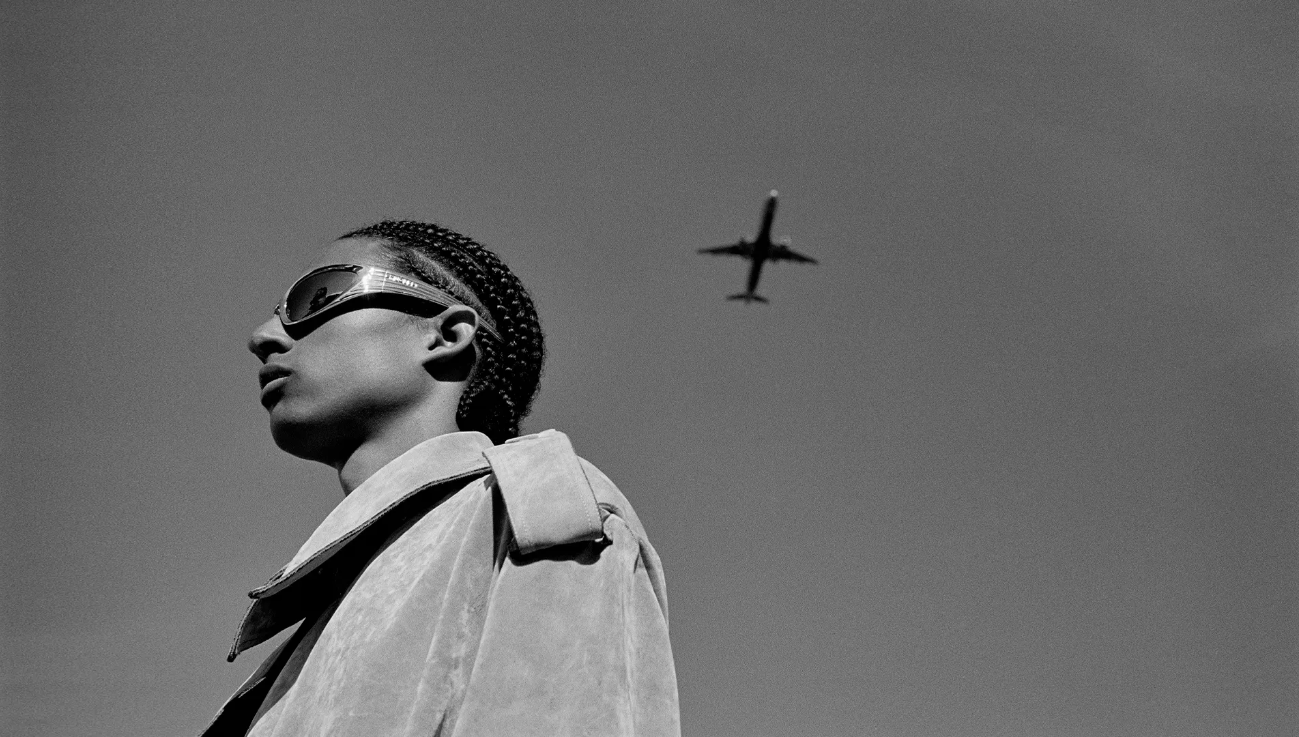 Black and white low-angle portrait of a person in sunglasses and coat with an airplane flying overhead