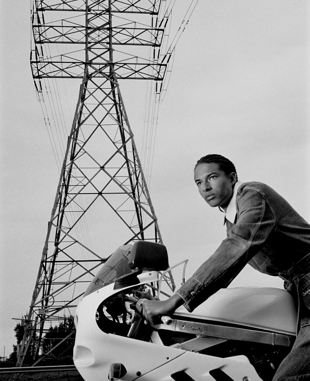 Monochrome portrait of a man on a motorcycle with an electrical tower, looking thoughtfully into the distance against a cloudy sky.