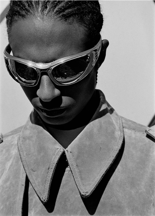 Monochrome portrait of a person with braided hair wearing futuristic sunglasses and a tan jacket against a light neutral background.