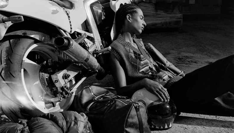 Black and white shot of a male with braids resting against a motorcycle with bags and helmet. Fashion editorial photography in studio.