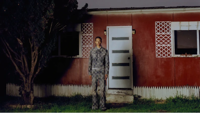 Man wearing a patterned denim jacket and matching jeans standing outside a red house at night