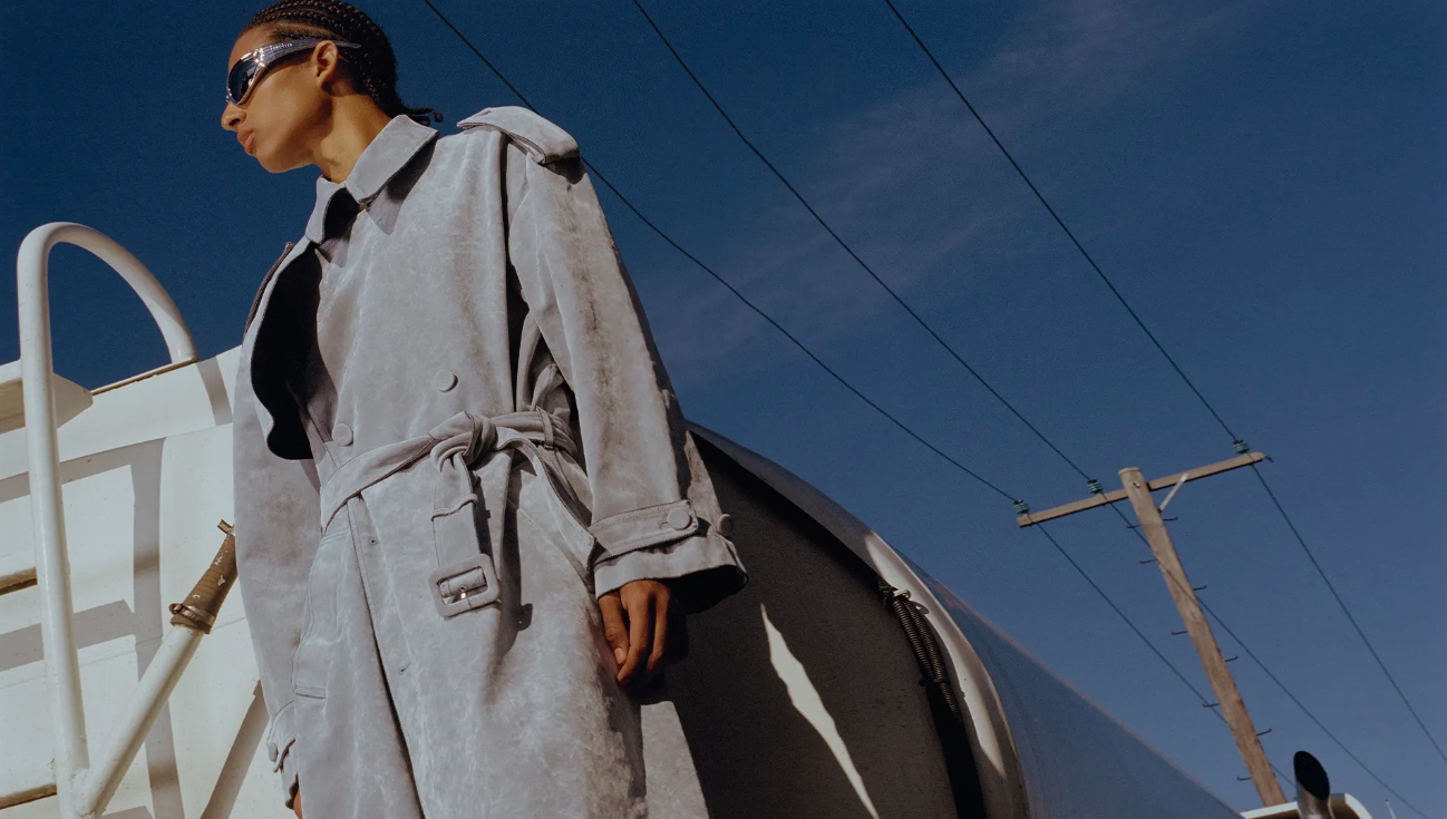 An adult fashion model wearing a grey trench coat and sunglasses stands on a white railcar against a bright blue sky with power lines.