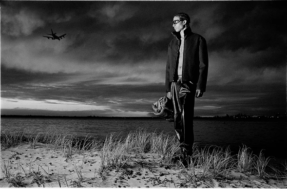 Adult male fashion model in jacket and leather pants poses on a beach under a cloudy sky with airplane overhead in a black and white photo.