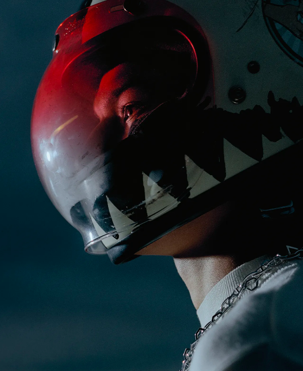 Close-up of an adult wearing a stylized helmet with an open faceplate. The person wears a chain necklace and a white collared shirt.