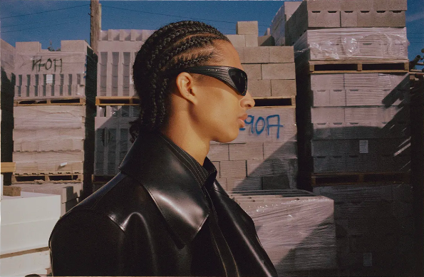 Adult with braided hair, wearing sunglasses and black coat in an outdoor setting with concrete blocks in the background.