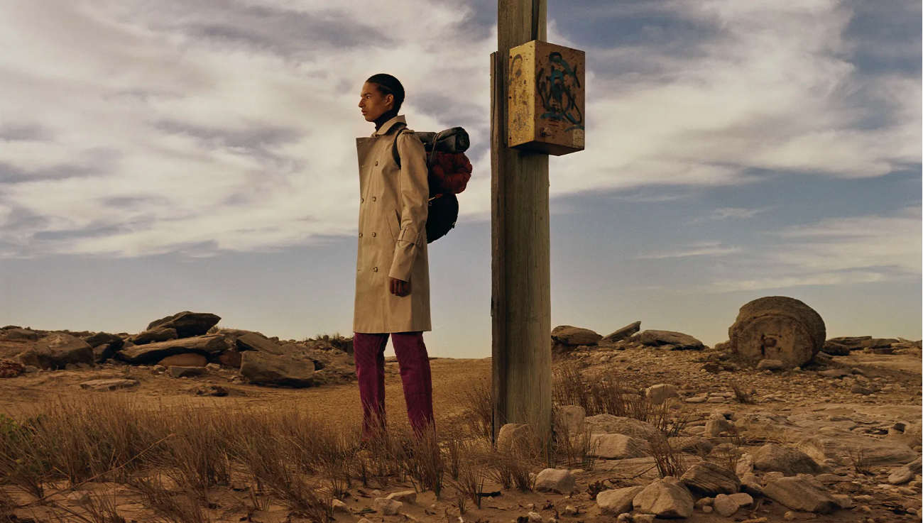 A slender adult fashion model stands on a rocky desert landscape, wearing a trench coat and carrying a backpack near a wooden post.