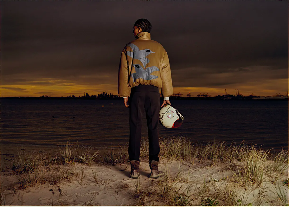 Man standing on a sandy beach holding a helmet, wearing a tan bomber jacket and dark trousers, facing a body of water at dusk.