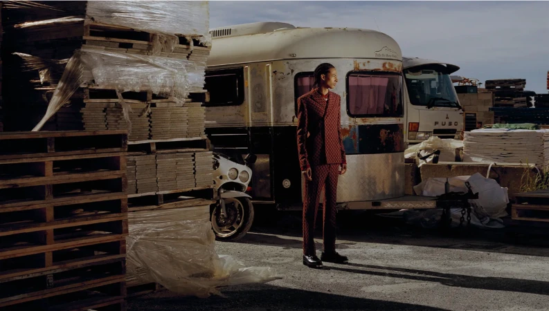 Adult man stands in front of an old trailer wearing a brown patterned suit on an outdoor industrial-looking lot with pallets.