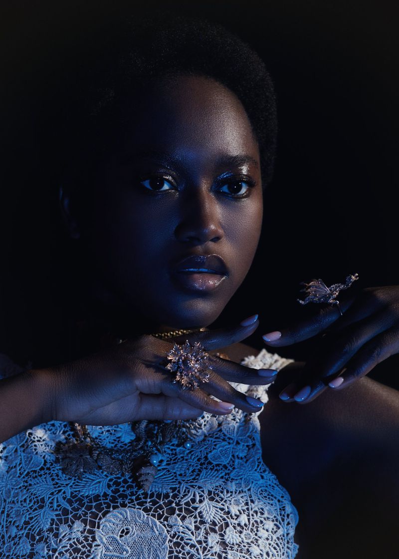 Portrait of a black woman with dramatic makeup and jewelry posing in a studio with dark, moody lighting against a black backdrop.