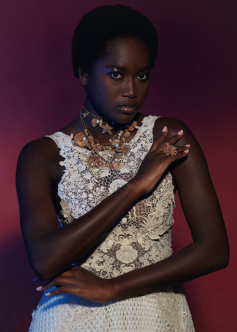 Portrait of a dark-skinned woman with a lace top and shell jewelry, posing in front of a red and blue backdrop in a studio setting.
