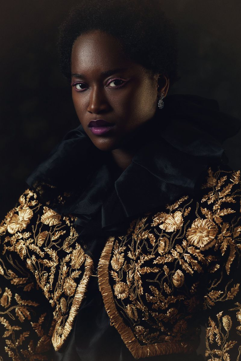 Portrait of an African American woman with dark hair, wearing a black velvet jacket with elaborate gold floral embroidery on a dark background.
