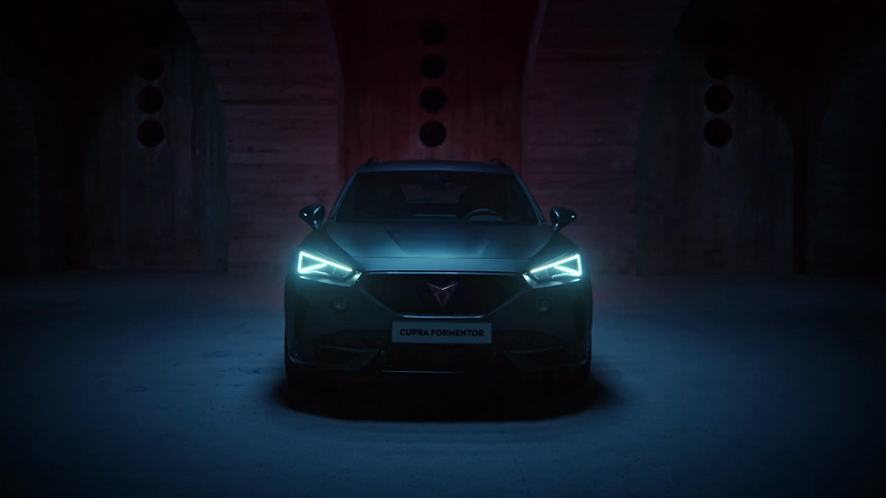 Front view of a dark gray Cupra Formentor car with bright headlights, positioned in a dimly lit garage or tunnel. Automotive imagery.