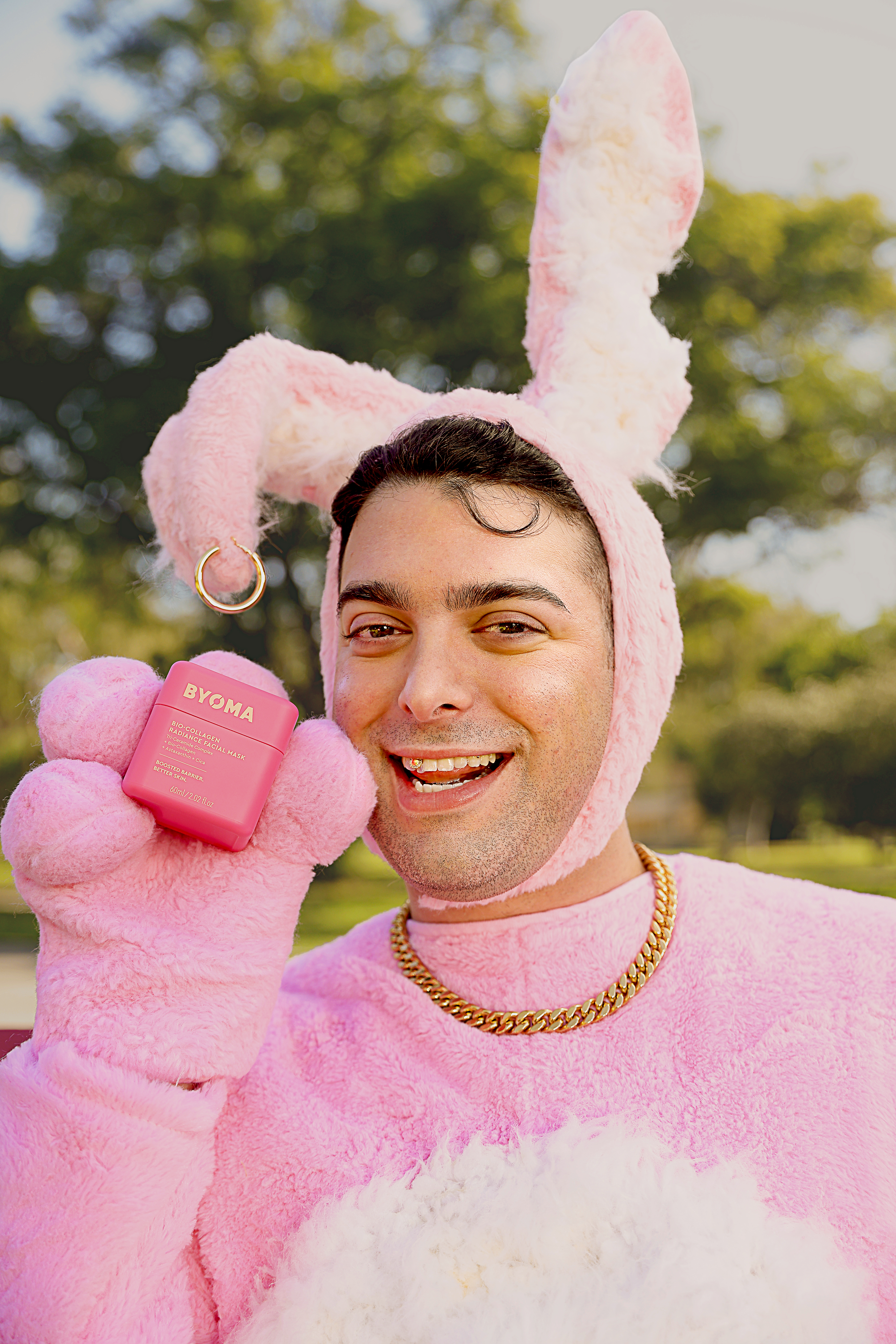 Adult male smiles wearing a pink bunny suit, holding a pink jar of face cream by Ryoma in an outdoor setting with greenery.