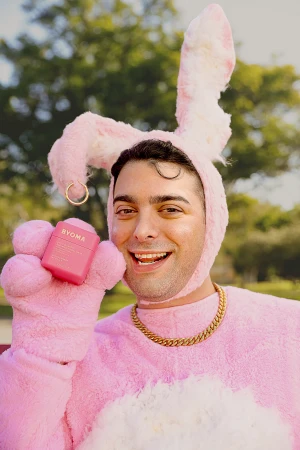 Adult male smiles wearing a pink bunny suit, holding a pink jar of face cream by Ryoma in an outdoor setting with greenery.