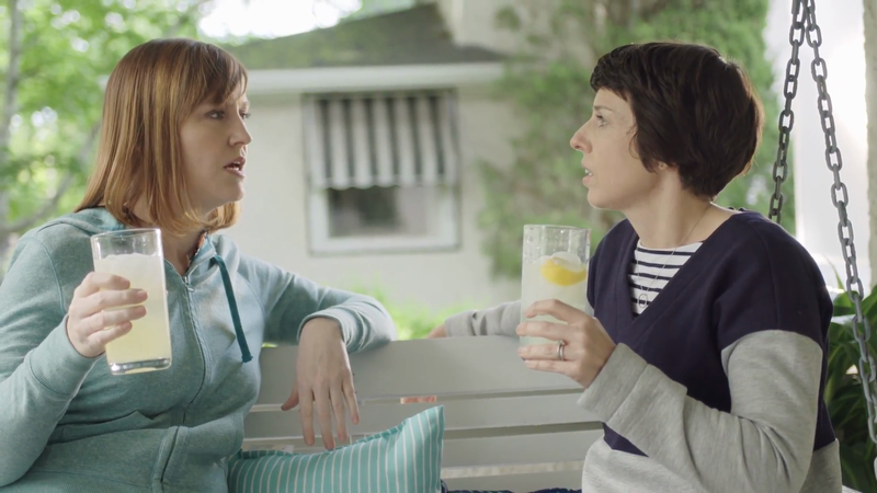 Two adult women on a white porch swing, talking and holding glasses of lemonade, with green foliage and a white house in the background.
