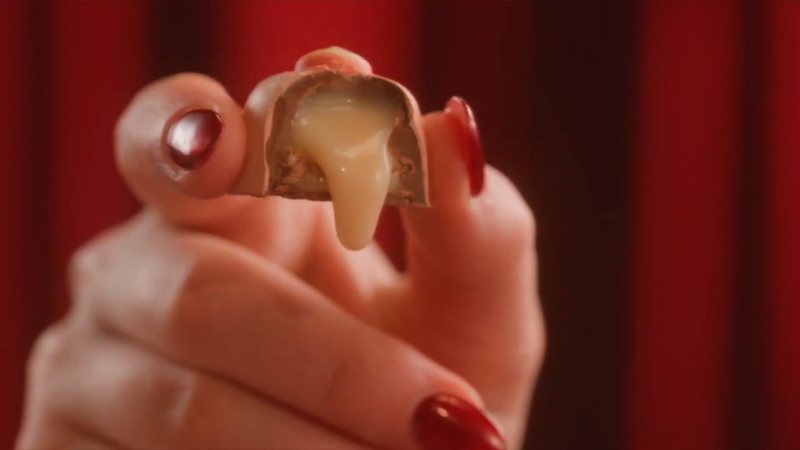 Close-up of a person holding a halved chocolate caramel candy, revealing the caramel center dripping downwards, against a red backdrop.