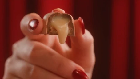 Close-up of a hand with red nail polish holding a bitten chocolate with flowing creamy filling against a red background.