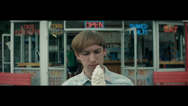 A young adult stands in front of a retro diner, enjoying a soft serve ice cream cone. The diner has vintage signage and a red bench.