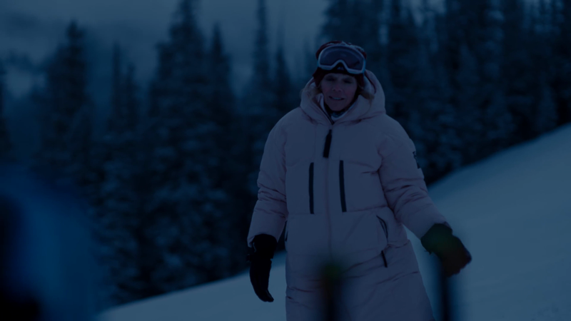 Woman wearing ski goggles and a winter suit stands on a snowy hill with pine trees in the background in a cold outdoor setting.