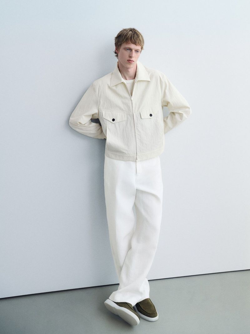 A blond male model in a casual pose. He wears an unzipped ivory jacket, white pants, and olive-green loafers against a pale background.