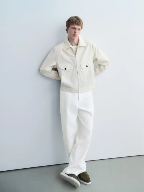A blond male model in a casual pose. He wears an unzipped ivory jacket, white pants, and olive-green loafers against a pale background.