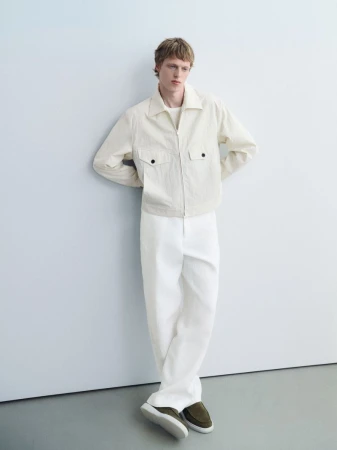 A blond male model in a casual pose. He wears an unzipped ivory jacket, white pants, and olive-green loafers against a pale background.