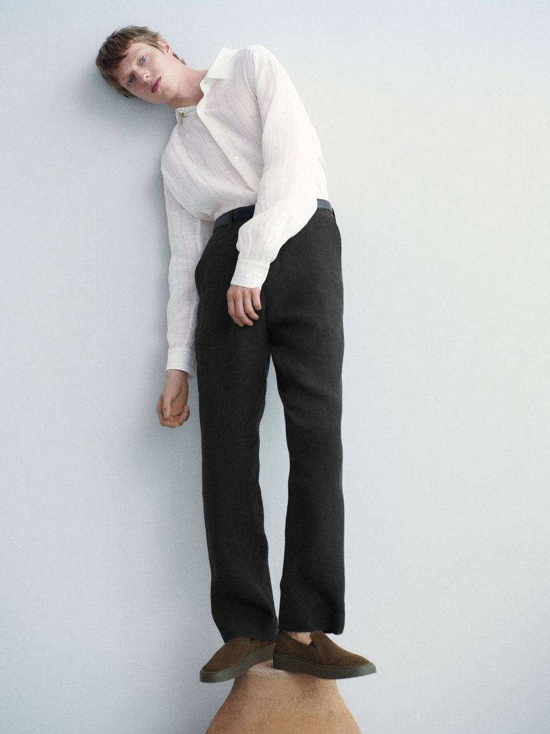 Adult male poses in a relaxed manner against a pale blue wall wearing a linen shirt, black pants, and olive shoes in a fashion editorial.