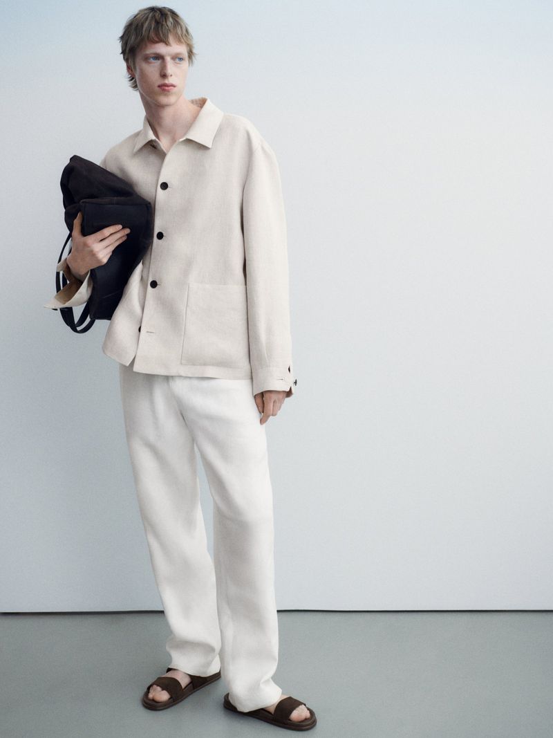 Fashion shot of a young man in an off-white jacket and pants, holding a black bag, standing against a pale blue-tinted white background.