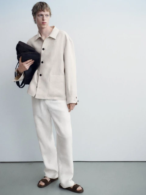 Fashion shot of a young man in an off-white jacket and pants, holding a black bag, standing against a pale blue-tinted white background.