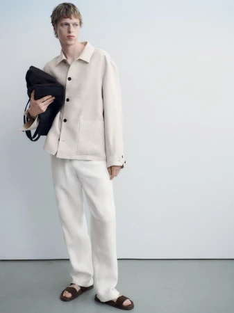 Fashion shot of a young man in an off-white jacket and pants, holding a black bag, standing against a pale blue-tinted white background.