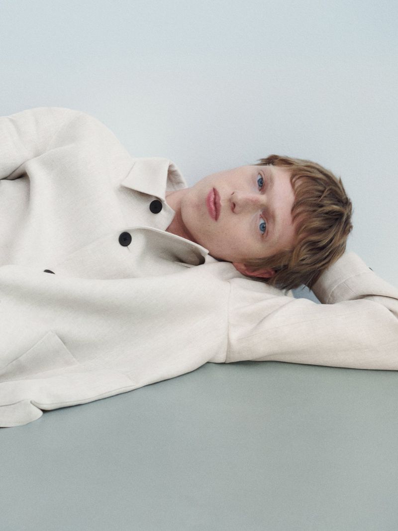 Portrait of a young adult with light skin lying down, wearing a light-colored blazer in a minimalist studio setting with blue background.