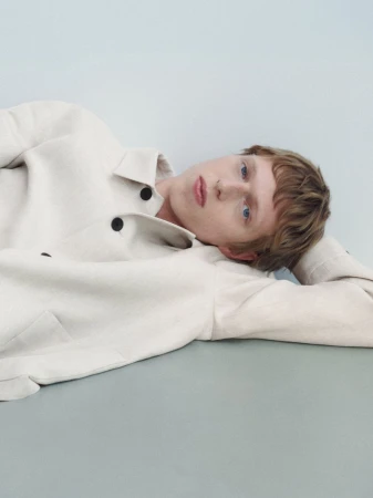 Portrait of a young adult with light skin lying down, wearing a light-colored blazer in a minimalist studio setting with blue background.
