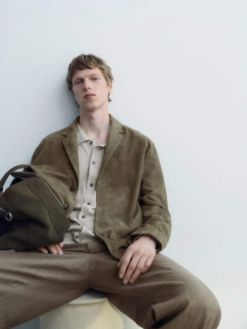 Adult man in olive jacket and beige pants, sitting, holding a bag, medium shot against a white wall. Casual fashion, indoor setting.