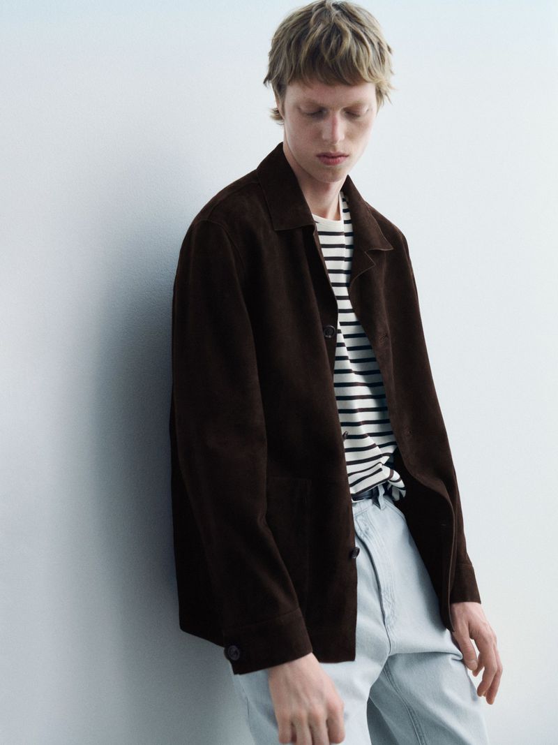 A fair-skinned male model wearing a brown jacket with a striped shirt stands against a white wall in a studio setting.