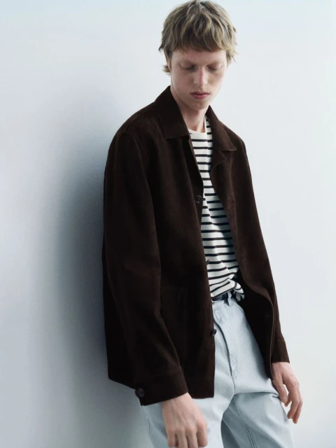 A fair-skinned male model wearing a brown jacket with a striped shirt stands against a white wall in a studio setting.